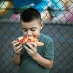 Chłopiec je kawałek pizzy pepperoni na tle siatki ogrodzeniowej