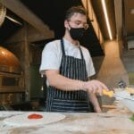 Kucharz w masce rozkłada składniki na cieście do pizzy w nowoczesnej kuchni