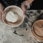 Ręce w rękawiczkach oprószają mąką surowe ciasto w bambusowym parowniku