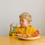 Uśmiechnięte dziecko przy stoliku popija napój i je pizzę